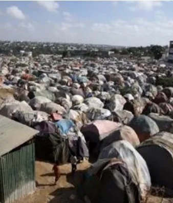 An arid, dusty refugee camp under a bright sky with people walking between tents
