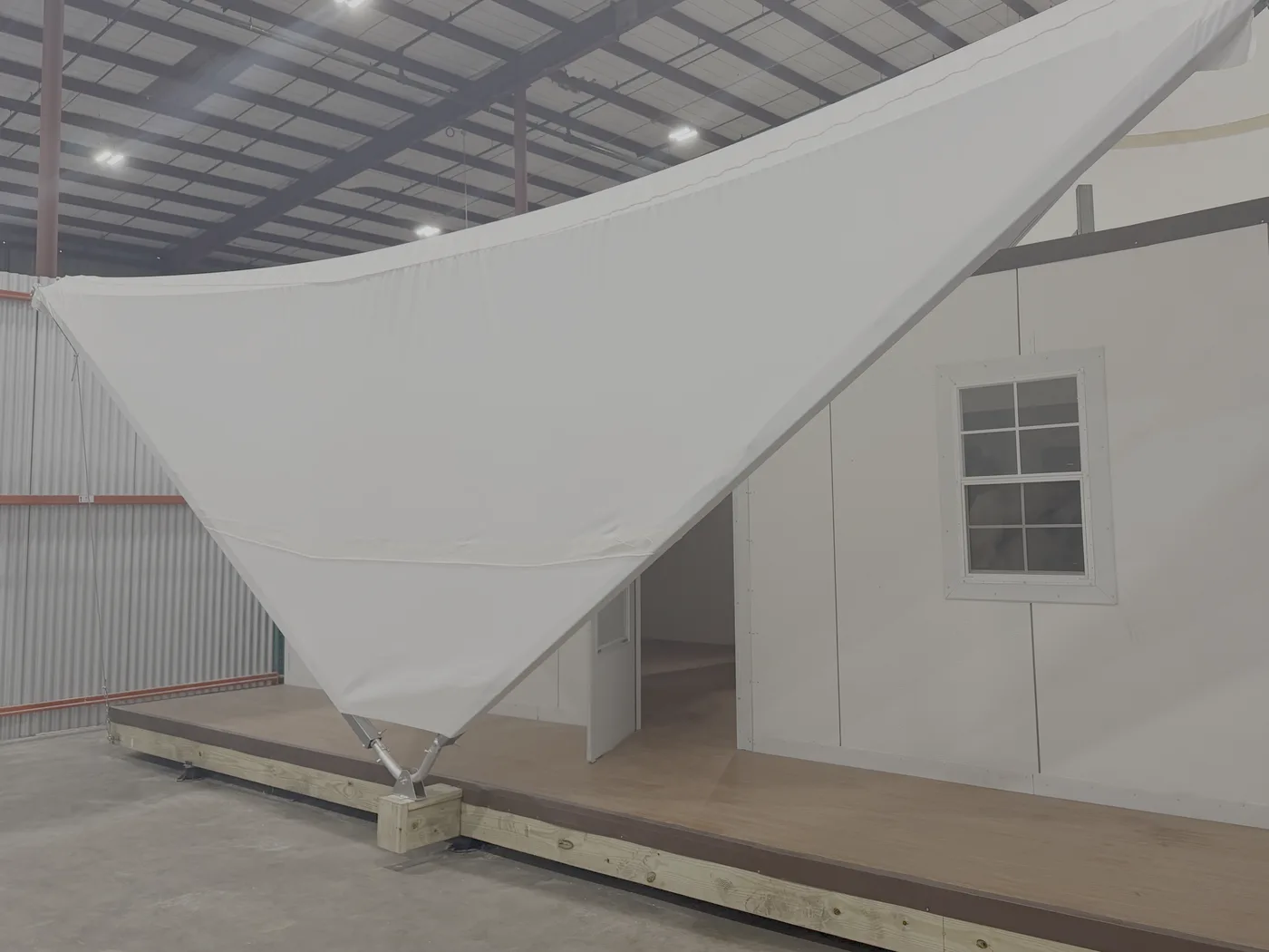 An Estia Homes unit assembled on a raised timber deck, with the fabric canopy tensioned overhead and anchored at the deck edge, and the panelled wall and doorway behind it